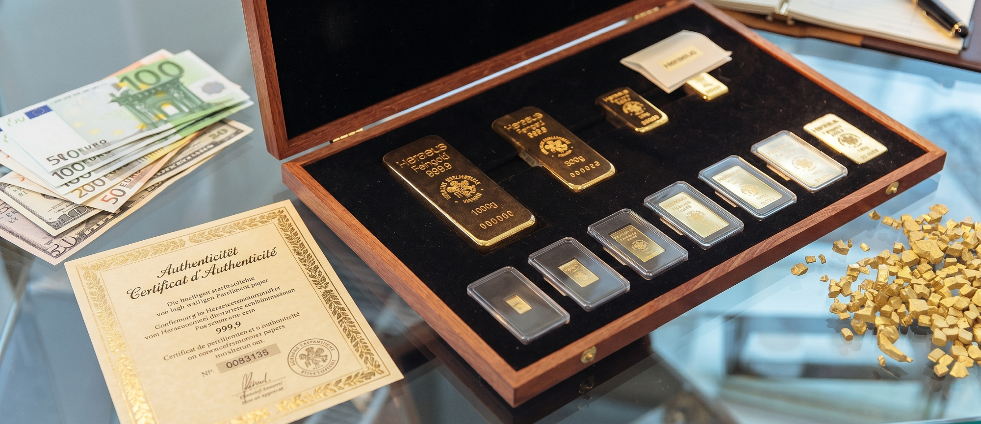 Goldbarren-Sammlung-mit-Zertifikat-und-Bargeld Eine geöffnete Holzschatulle mit verschiedenen Goldbarren, ein Echtheitszertifikat und Euro-Geldscheine auf einem Glastisch.